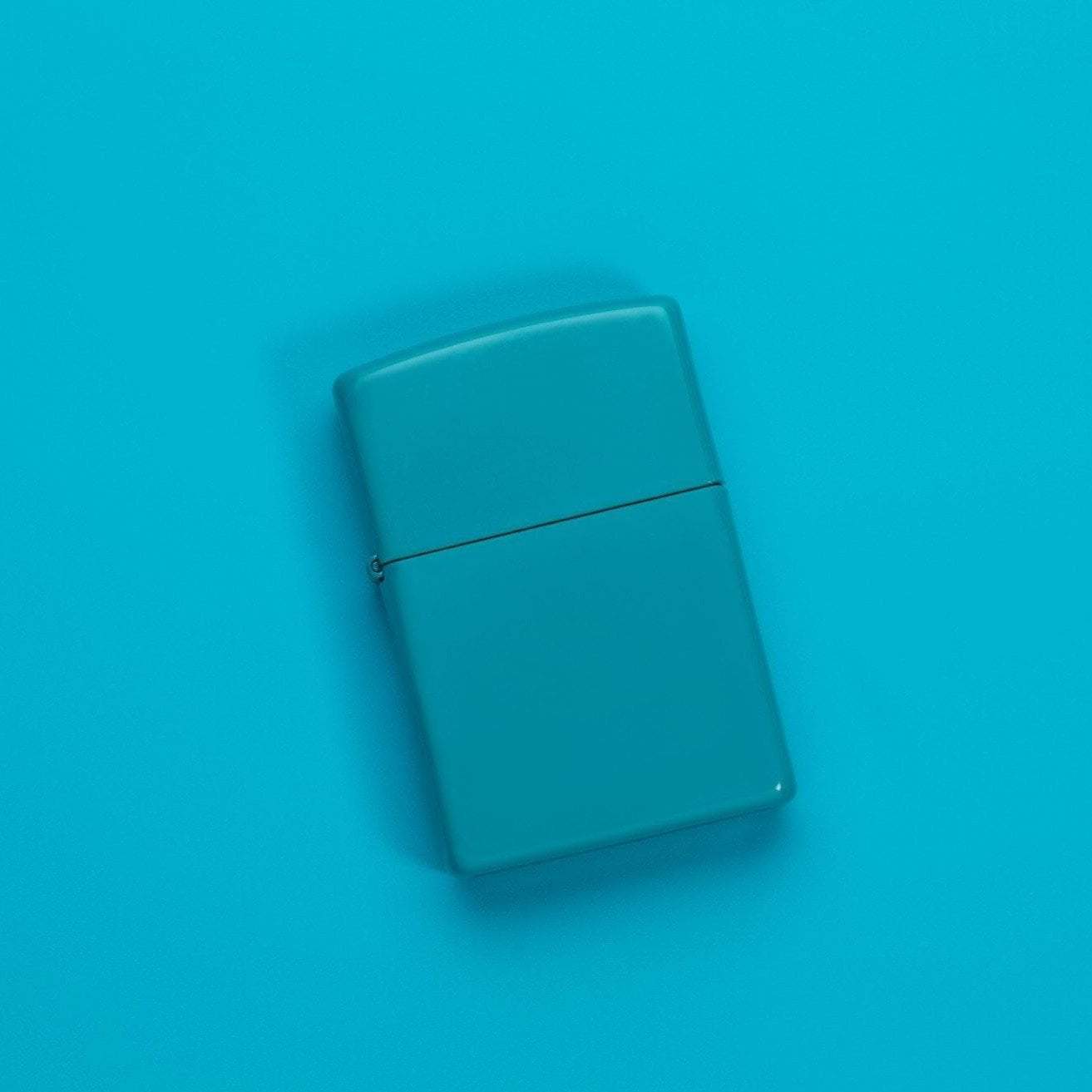 Lifestyle image of Classic Flat Turquoise Windproof Lighter laying on a turquoise surface